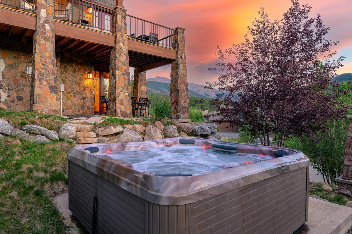 An inviting exterior scene featuring a private hot tub on a stone patio, surrounded by the home's stone façade. Comfortable balcony seating is positioned to enjoy sweeping sunset views over the mountains, creating a luxurious and tranquil setting for group relaxation and socializing.