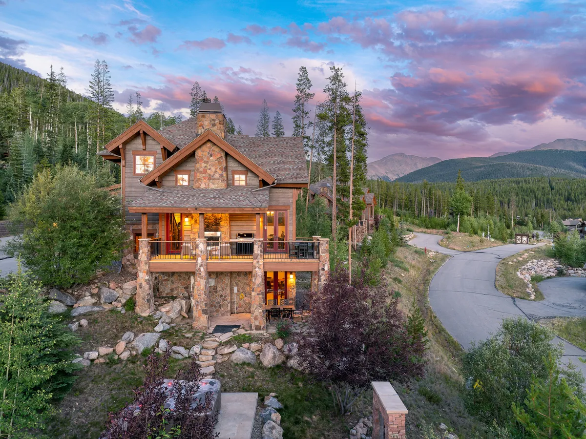 A tranquil mountain home exterior with a stone facade, a welcoming porch with seating, expansive windows, and lush surroundings. The setting evokes relaxation, outdoor socializing, and quick access to nearby hiking trails or nature walks.