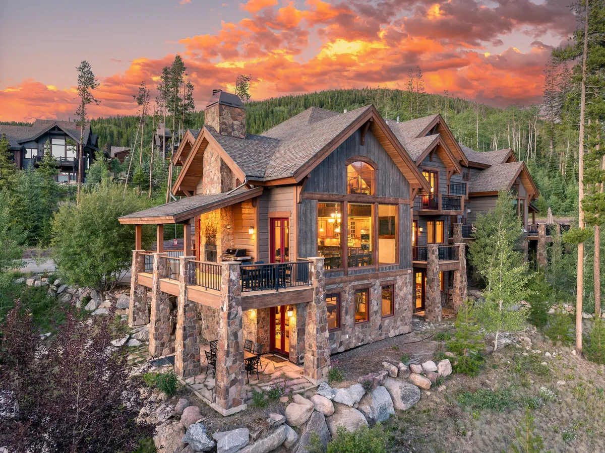 The hero image showcases the impressive stone-pillared entrance of a luxury villa, with expansive windows glowing warmly from within. The house is nestled in a tranquil natural landscape, with balconies perfect for gatherings, a cozy fire pit area, and breathtaking mountain views, all framed by the soft hues of a Colorado sunset.