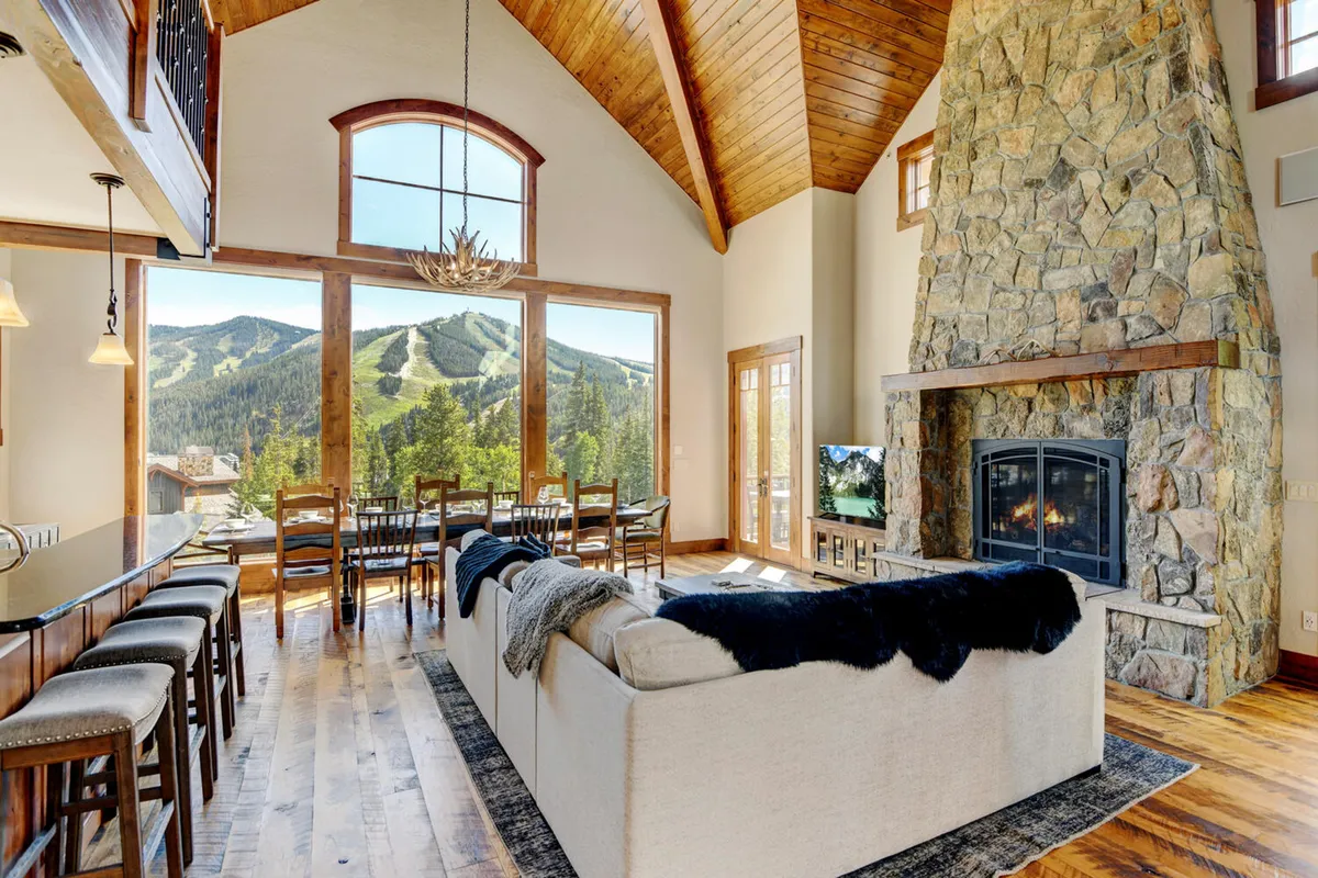The image showcases a cozy living area with a prominent large stone fireplace, expansive windows offering dramatic mountain views, a dining area beside the windows, and a comfortable sofa. The ambiance is warm and welcoming, with touches that evoke both comfort and a connection to the outdoors.