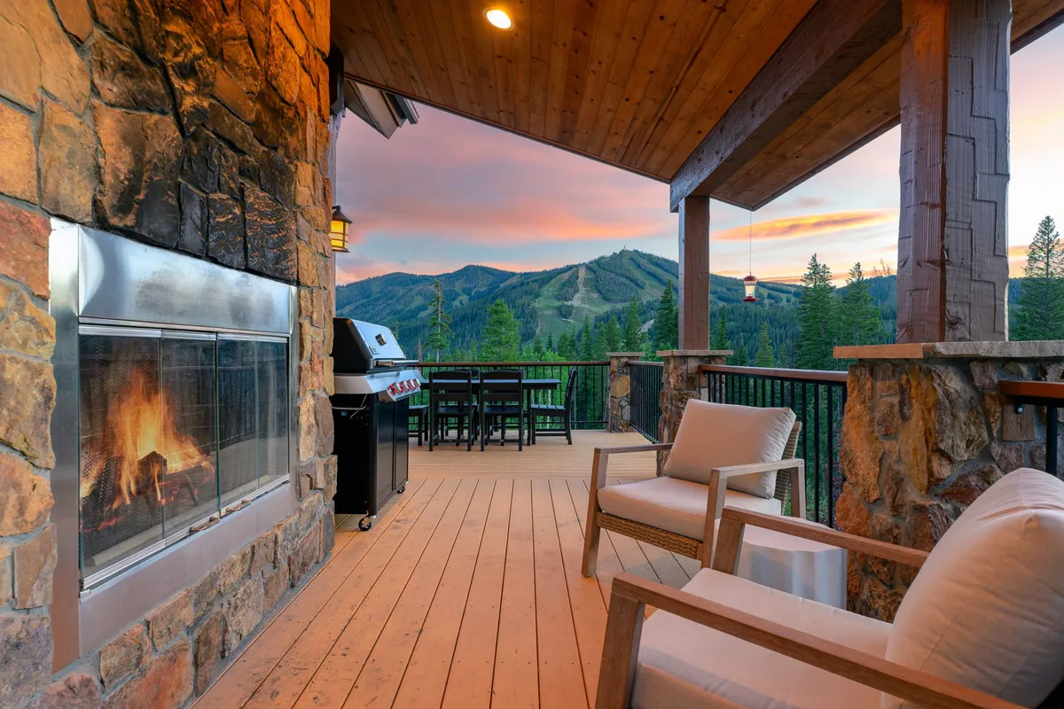 An expansive main deck looks out across the mountains, featuring cozy fireside seating, a natural gas grill, and a dining table for outdoor gatherings. The scene is set for both sunrise coffees and après-ski evenings, with the warm glow of the fire contrasting against the tranquil mountain backdrop.