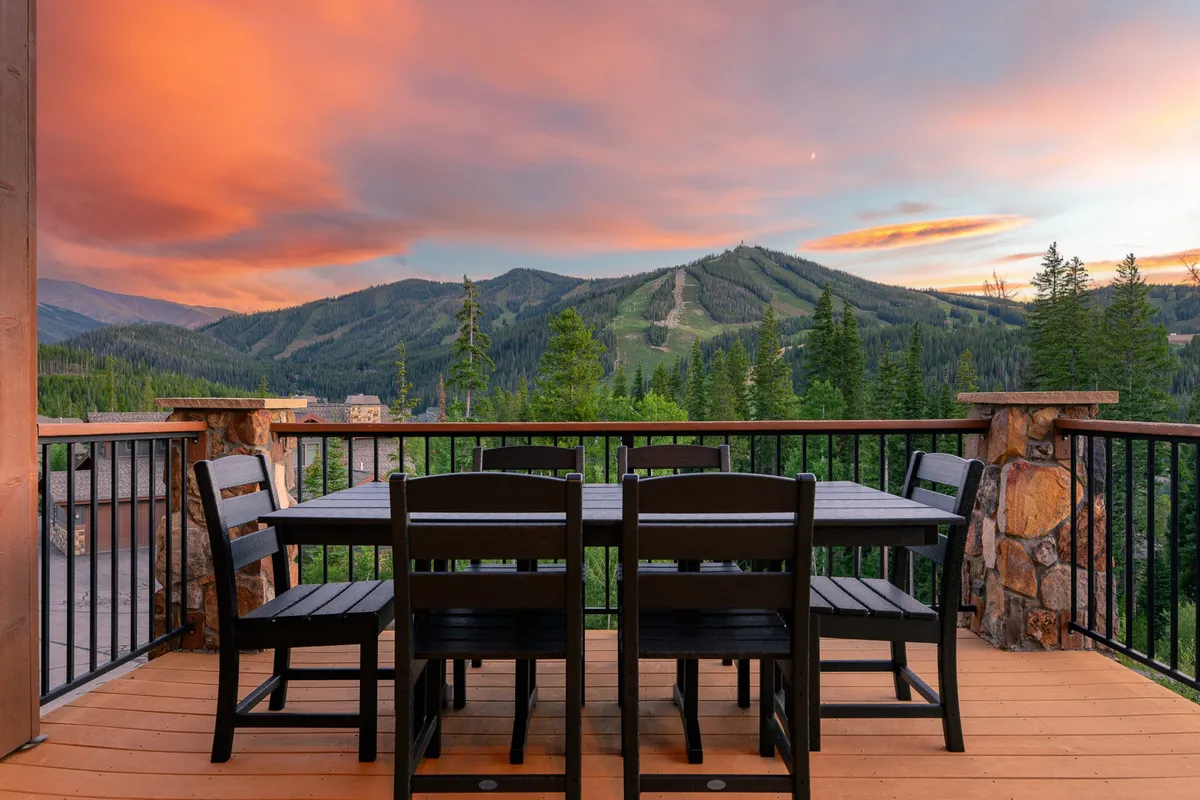 A serene outdoor dining area with a large table set for a group, surrounded by breathtaking mountain views and illuminated by the warm glow of a sunset sky. The atmosphere suggests shared meals, conversations, and group gatherings in nature.