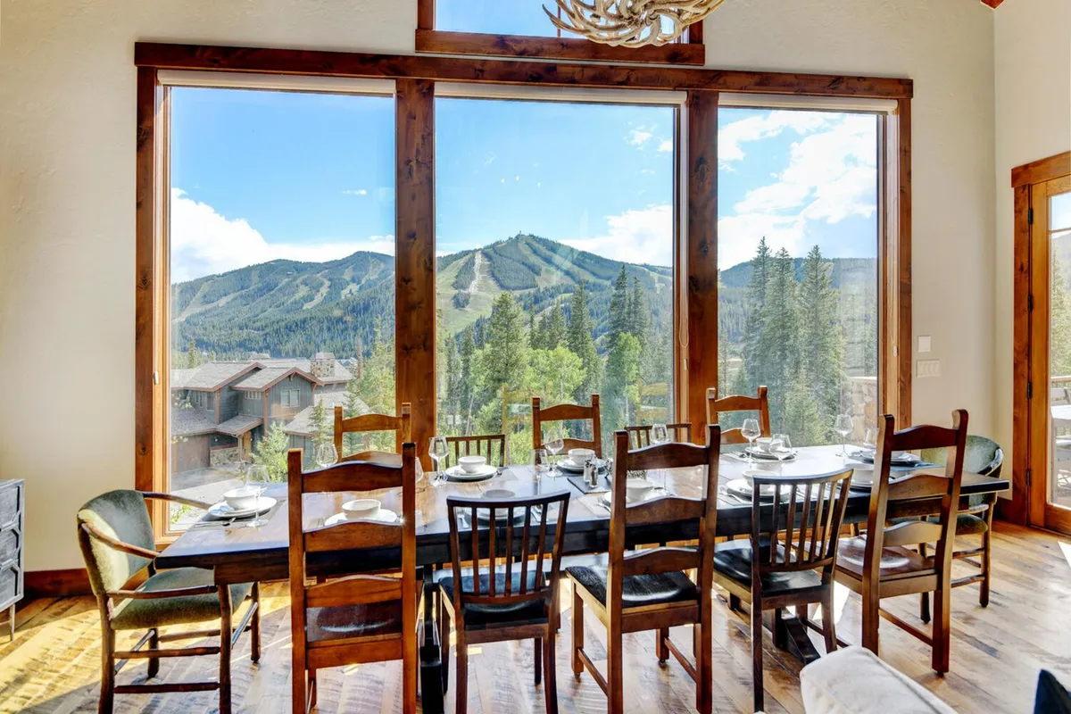 A large wooden dining table is set beside expansive windows with a mountain view. Wooden beams and a statement chandelier above create a cozy, communal atmosphere, ideal for family feasts or casual gatherings and game nights.