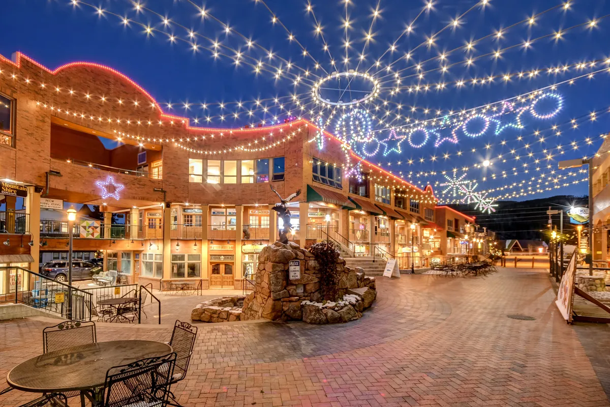 A festive village square at dusk, featuring decorative lights, star-shaped ornaments, a cozy stone fountain, and outdoor seating. The scene is alive with people enjoying food, shopping, and gathering for community events.