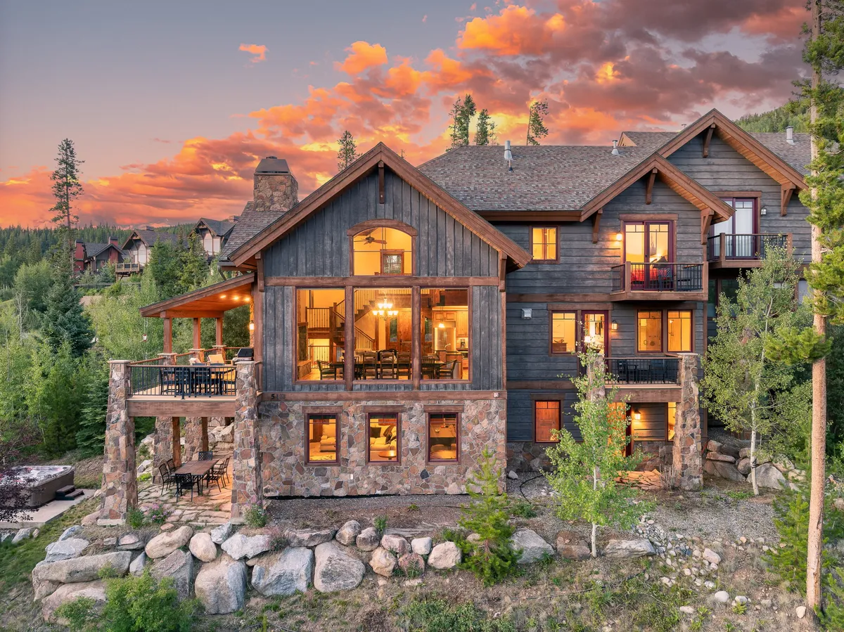 An inviting outdoor living space featuring a wooden balcony with large glass windows, a stone facade, and comfortable seating. The area is surrounded by natural beauty, offering a vantage point for enjoying sunsets, stargazing, or gathering by a fire pit with mountain views beyond.