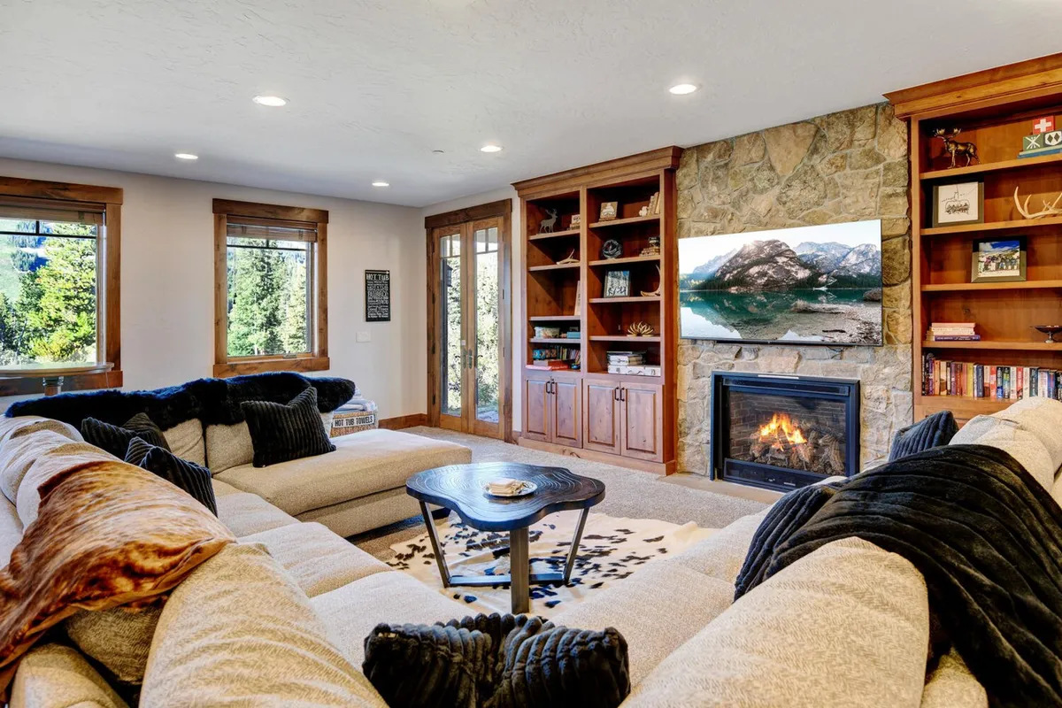 A spacious living room with a large sectional sofa faces a welcoming stone fireplace. Expansive windows frame panoramic mountain views, while a 55" Smart TV is positioned above the fireplace. Wooden bookshelves line one wall, and the room is bathed in warm, natural light. The decor blends contemporary comfort with mountain charm, creating an inviting atmosphere for group gatherings.
