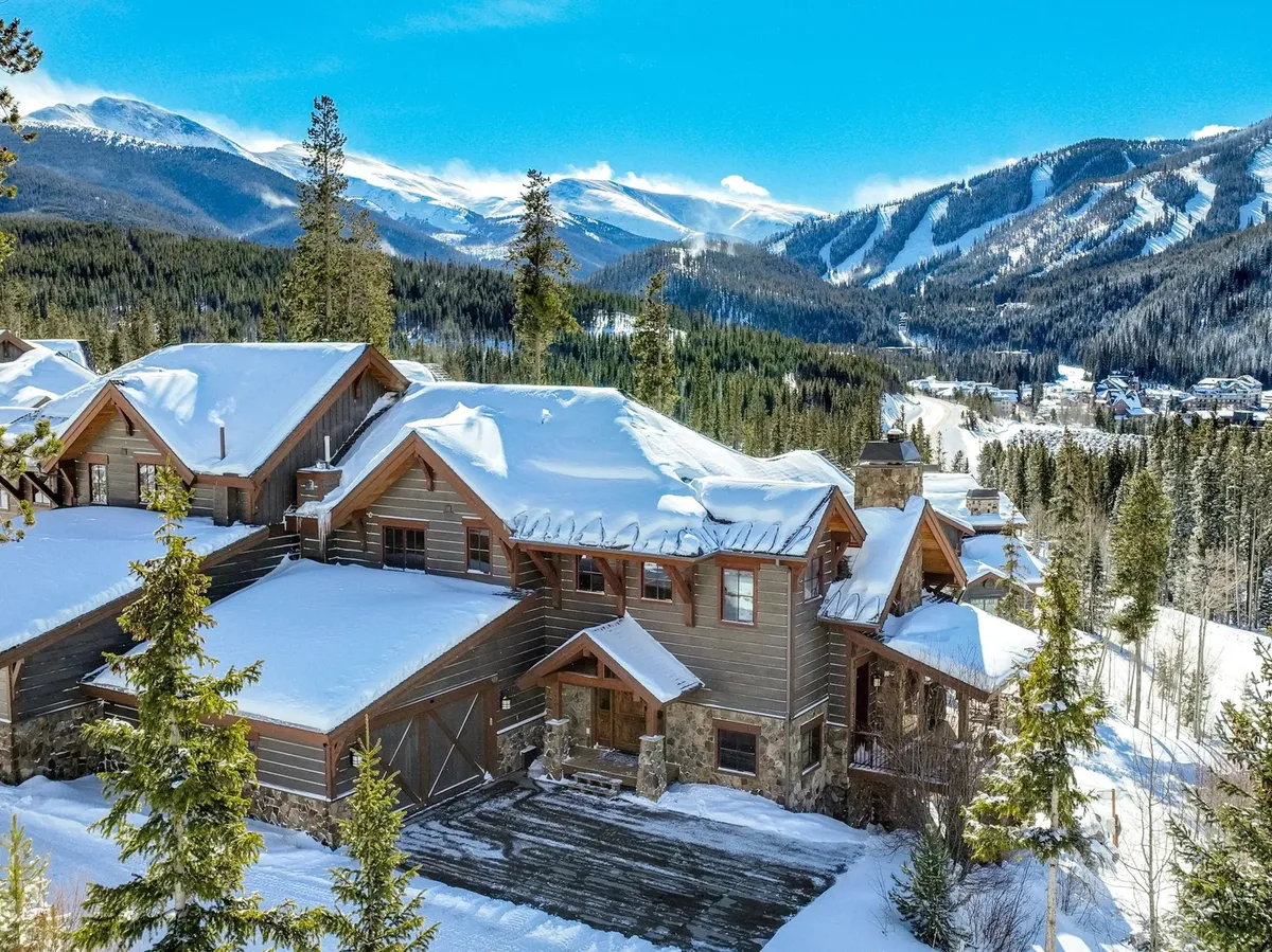 A snow-covered luxury villa with a wooden exterior and stone accents, nestled among pine trees. The early morning light casts a pink glow over the roof, while the majestic mountain backdrop hints at proximity to winter sports. The setting exudes a mix of adventure and coziness, perfectly reflecting mountain living.